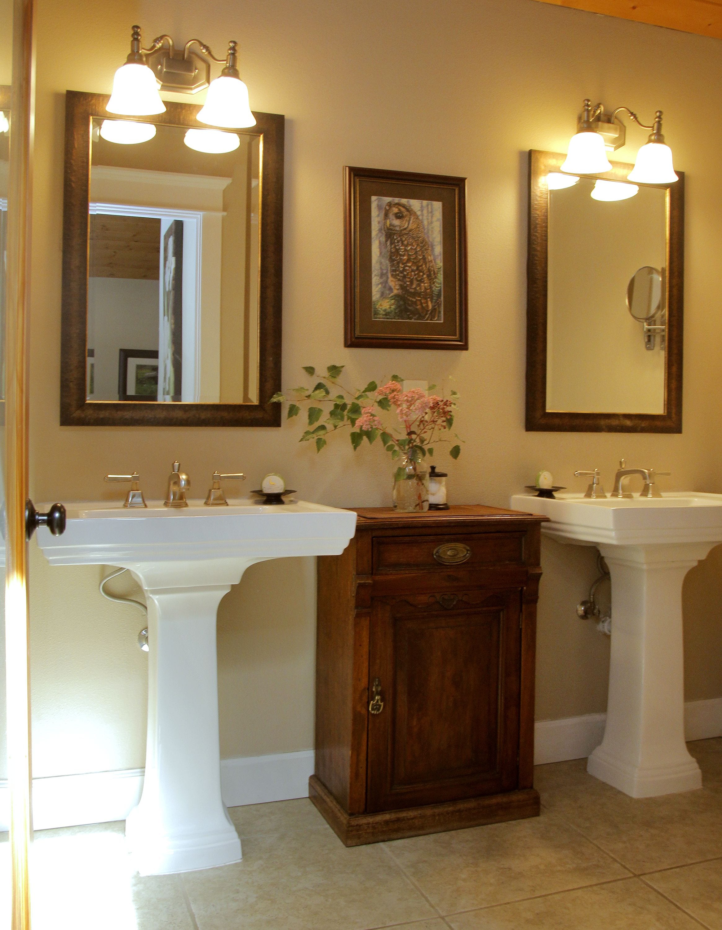 A bathroom with two white pedestal sinks, a wooden cabinet, and two wall-mounted mirrors with light fixtures.