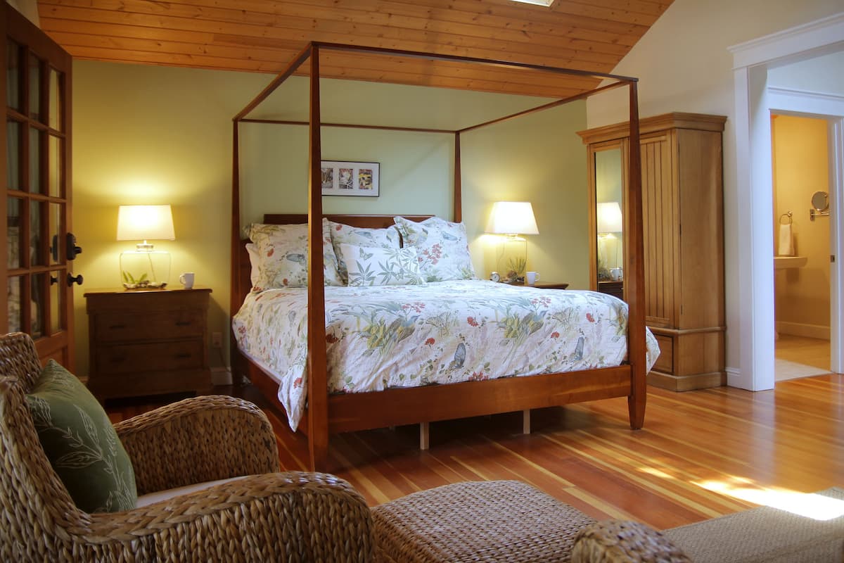 A four-poster bed with floral bedding, a wooden floor, wicker furniture, and a wooden ceiling.