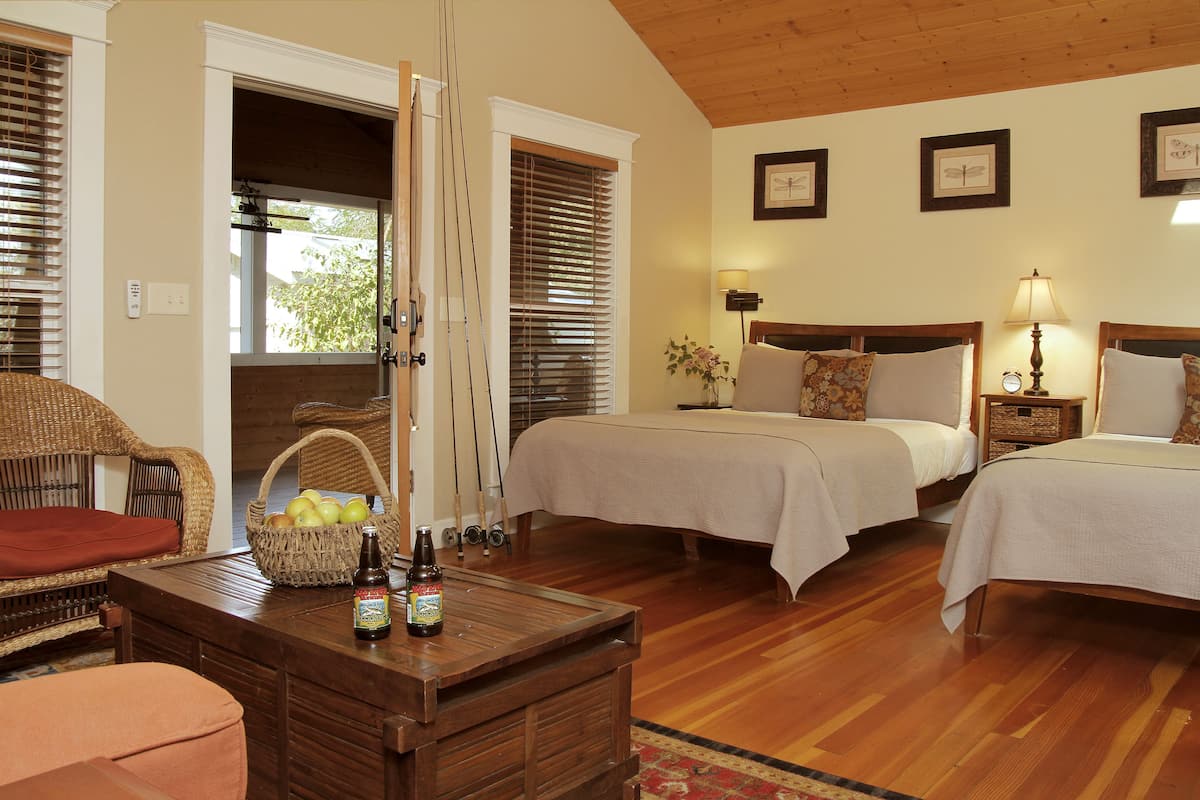 A hotel room with two beds, a wicker chair, a wooden table with bottles, and a basket of apples.
