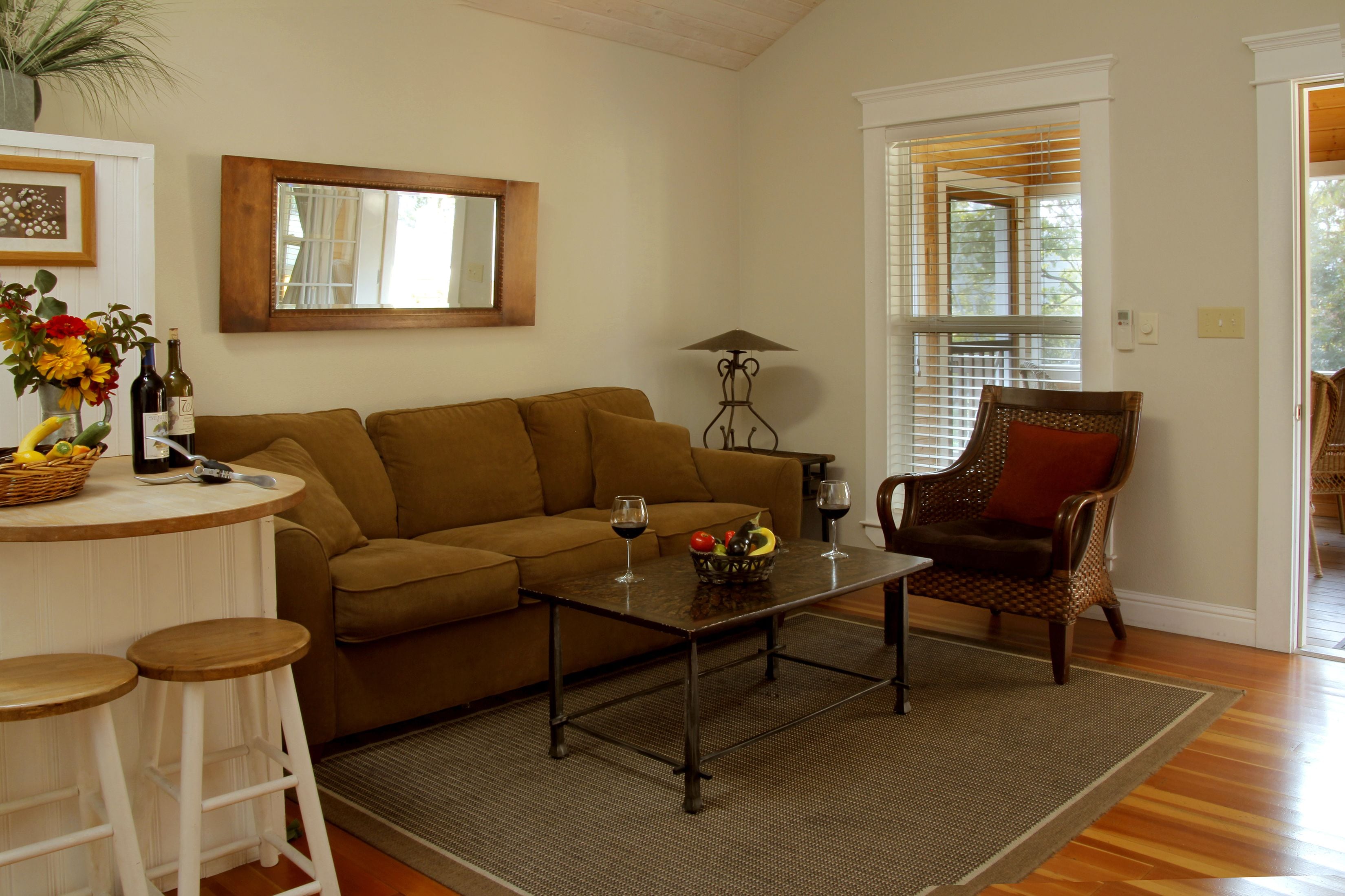 A living room with a brown sofa, a coffee table with wine glasses, a wicker chair, and a dining area with stools.