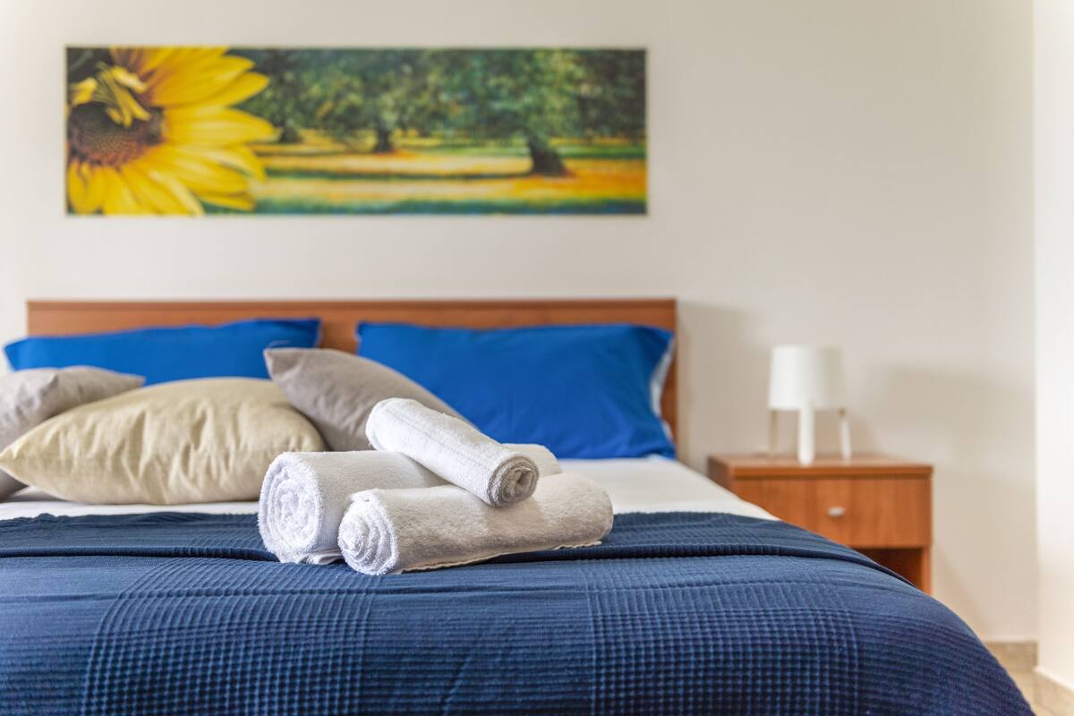 A neatly made bed with blue and beige pillows, a blue bedspread, and rolled towels on the bed.