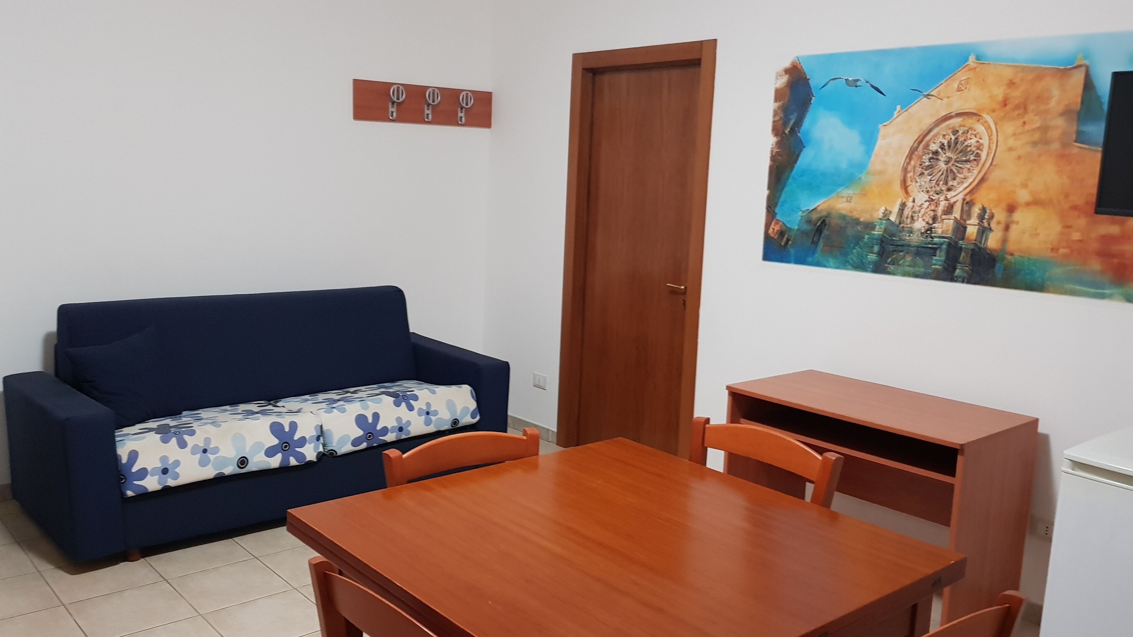 A compact living space with a sofa, a dining table, and chairs. A door is visible, and a painting hangs on the wall.