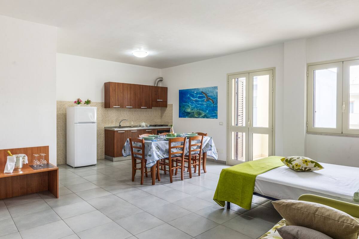 A compact kitchen with wooden cabinets, a dining table with chairs, a bed with a green blanket, and a painting of an underwater scene.
