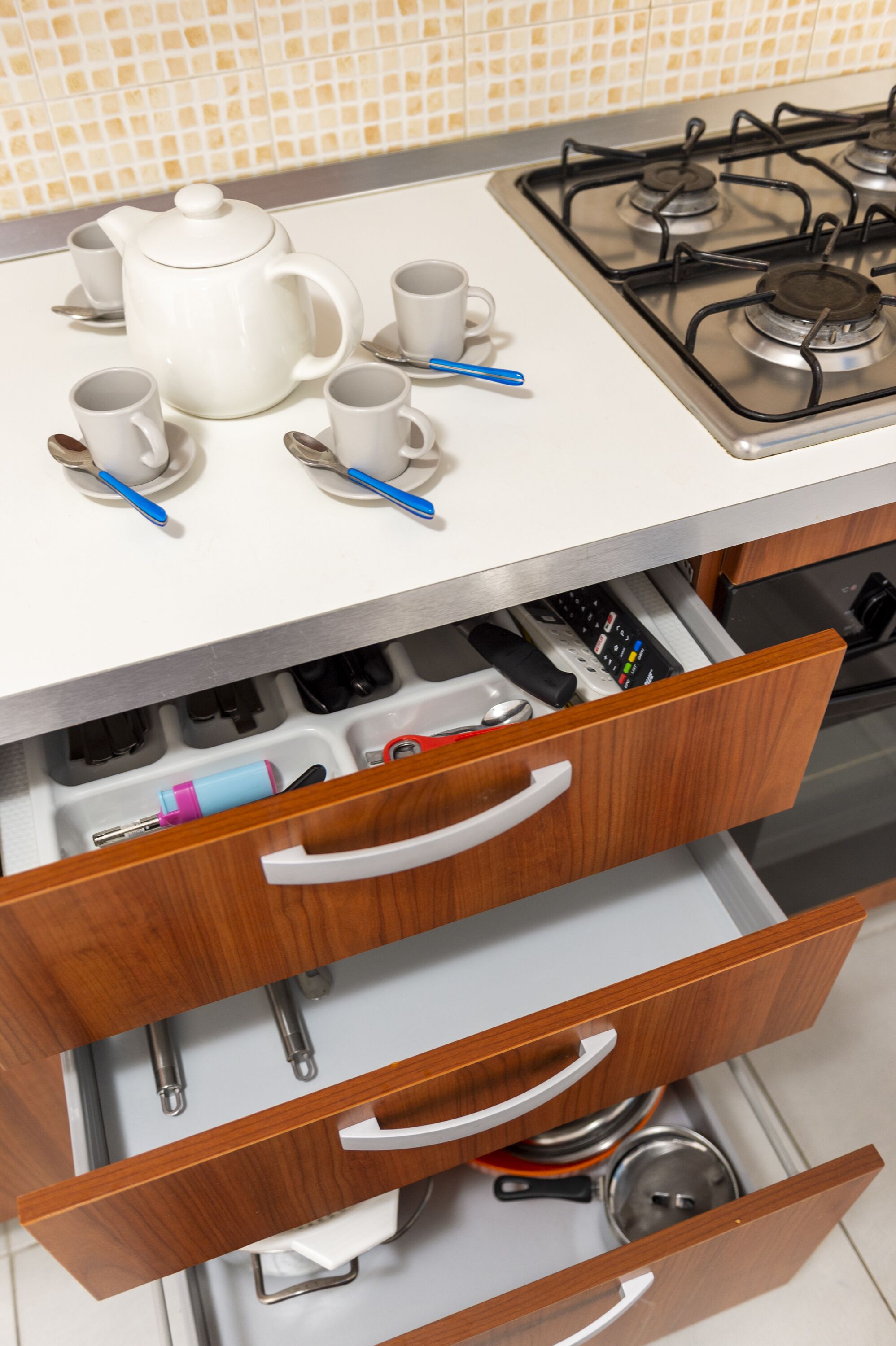 Una cocina con estufa a gas, tetera blanca y tazas y cucharas a juego. Abajo, hay cajones de madera con utensilios y elementos de cocina.