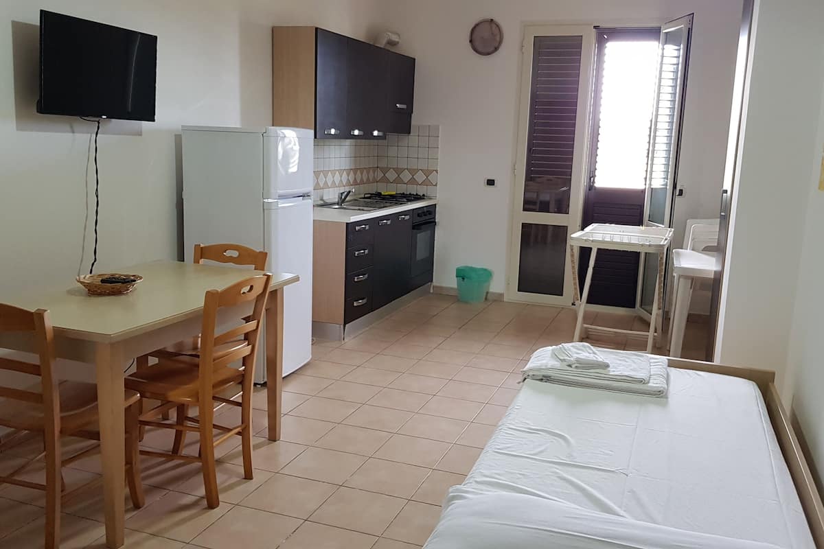 A compact kitchen with a dining table, chairs, a refrigerator, a stove, and a sink. There is a small balcony with a chair and a drying rack.