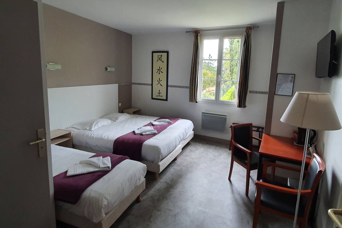 A hotel room with two beds, a table with chairs, a window with curtains, a radiator, and a wall-mounted TV.
