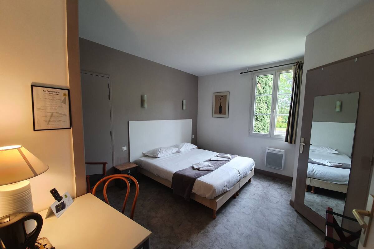 A hotel room with a bed, a desk, a chair, a lamp, a window, and a mirror.