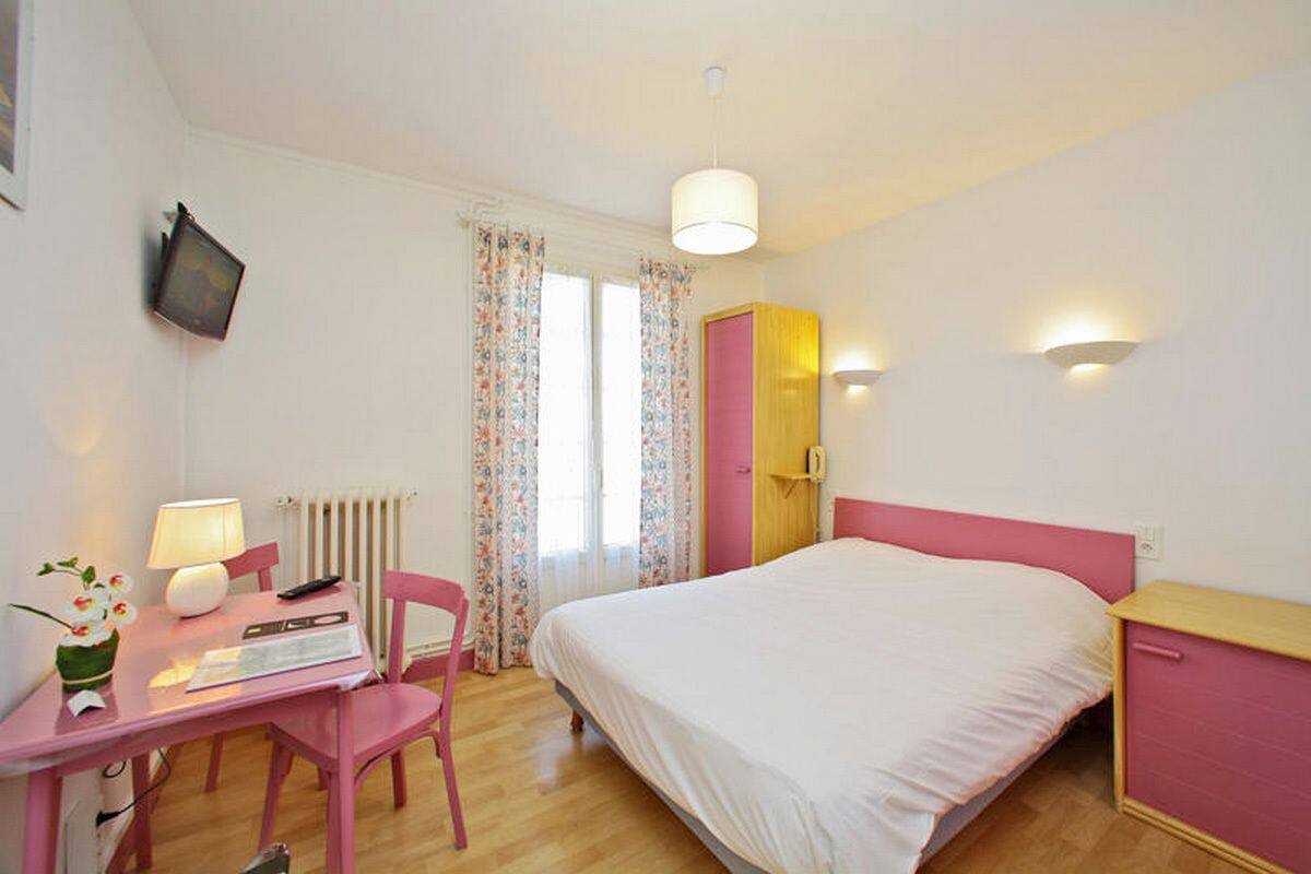 Een slaapkamer met een bed, een bureau, een stoel, een lamp, een radiator, een televisie en een raam met gordijnen.