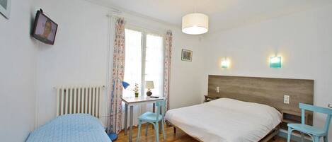A bedroom with a wooden bed, a desk with a chair, a radiator, and a window with curtains.