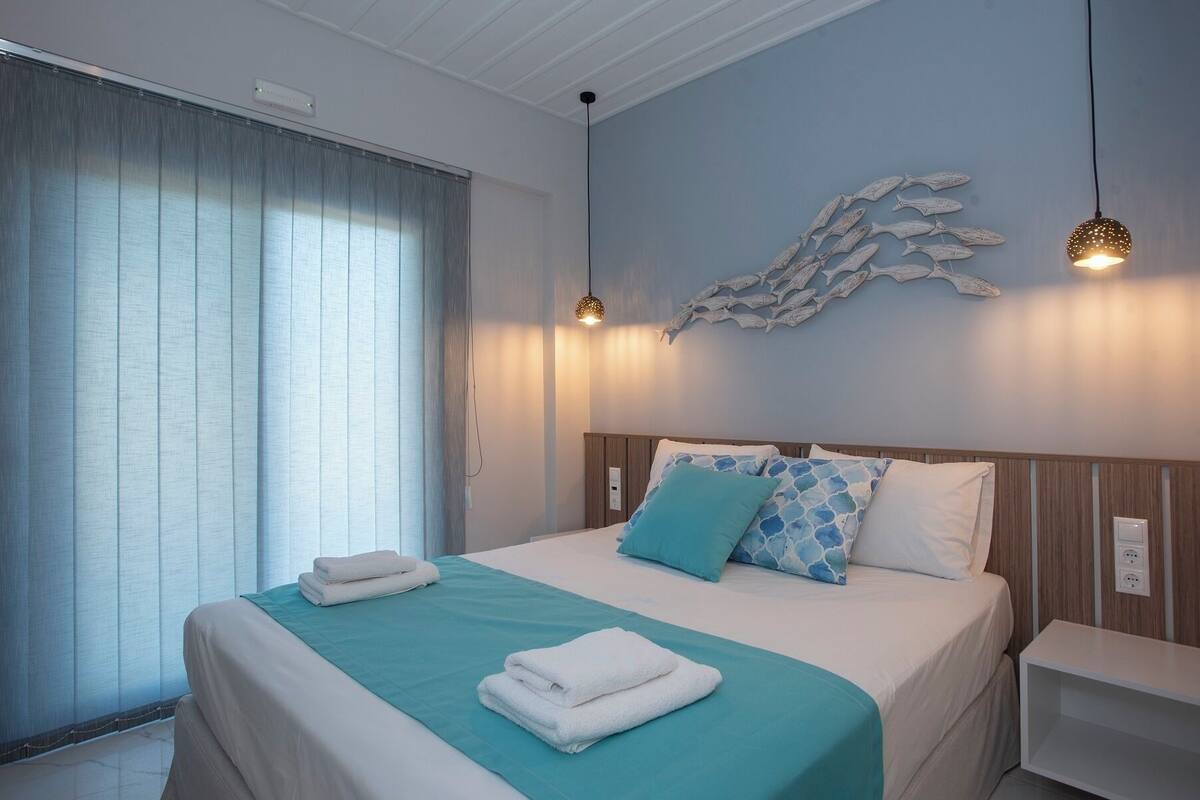 Uma cama bem arrumada com colcha verde-água e almofadas brancas, luminária fixada na parede e um quadro decorativo de peixes.