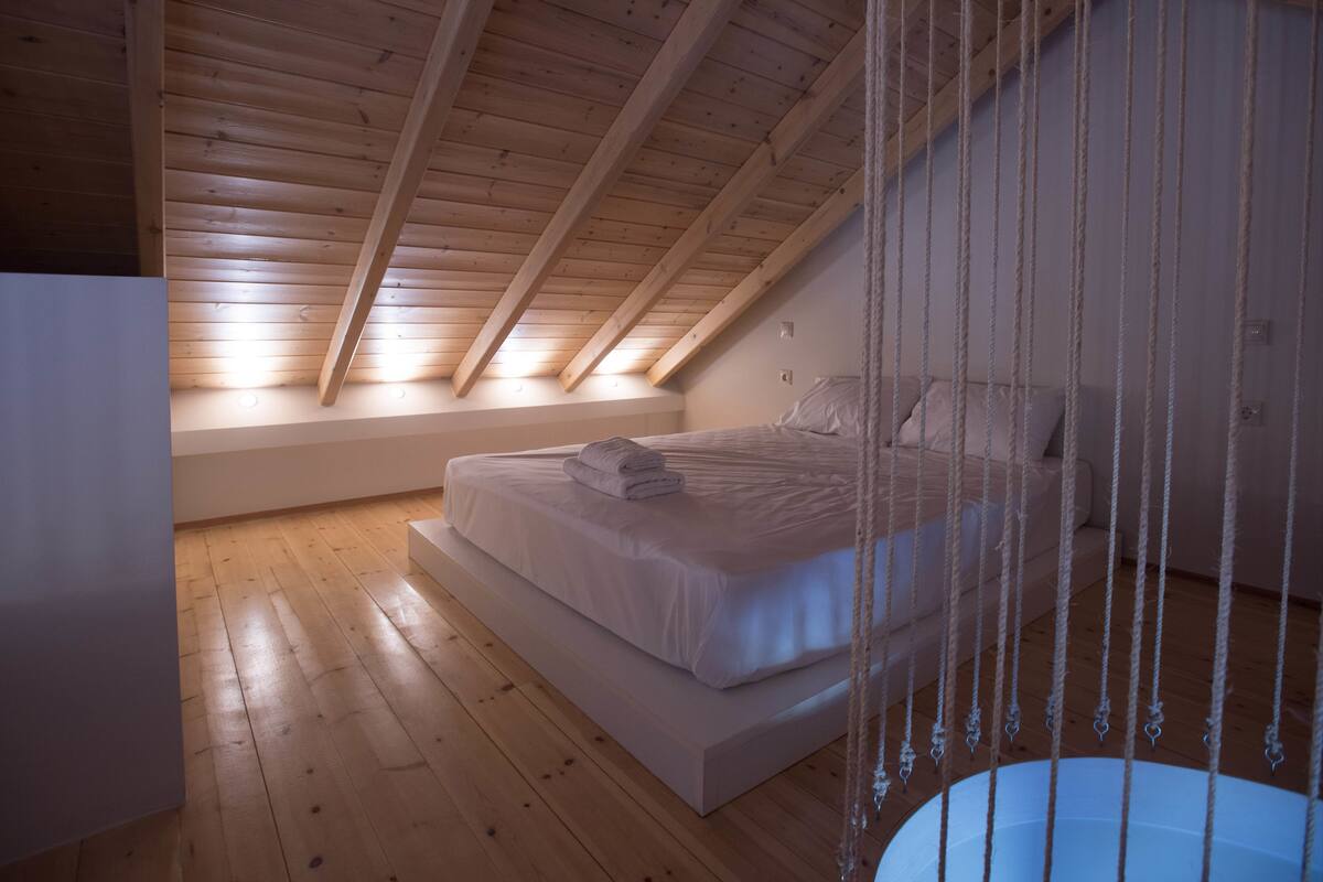 Quarto com teto inclinado, vigas de madeira, cama com roupa de cama branca e um balanço de corda pendurado.