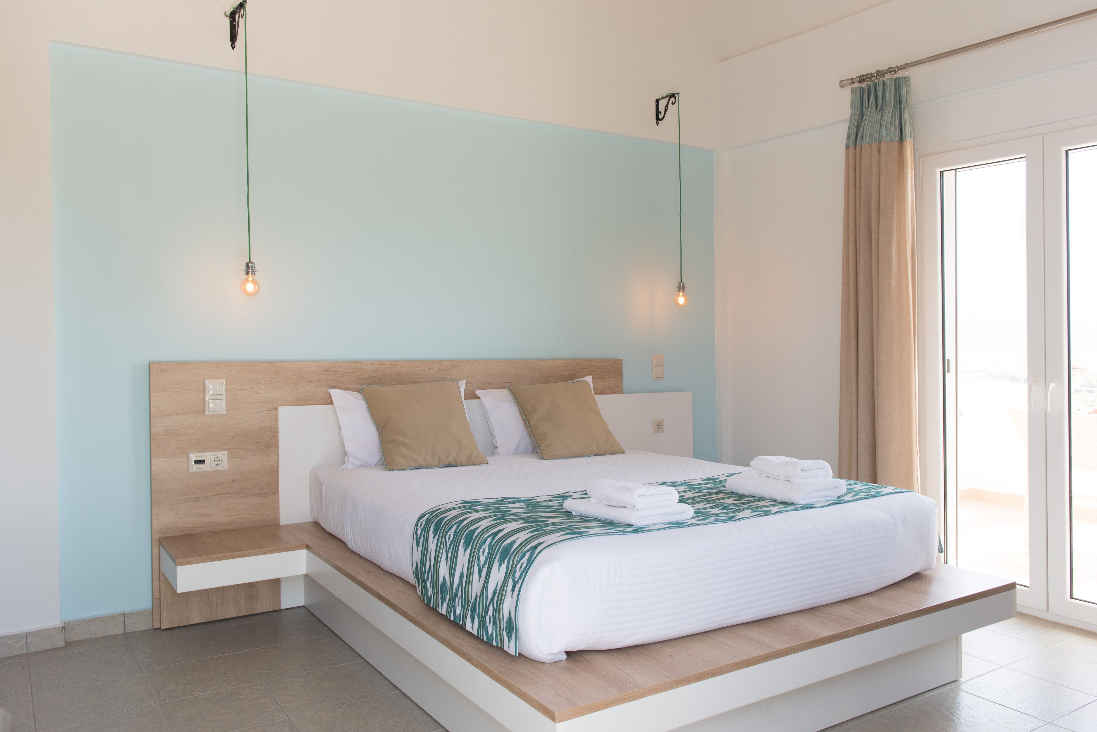 Ein modernes Schlafzimmer mit einem hölzernen Kopfende, einem Bett mit weißer Bettwäsche und einer grünen Tagesdecke mit Blätermuster, sowie zwei Pendelleuchten.