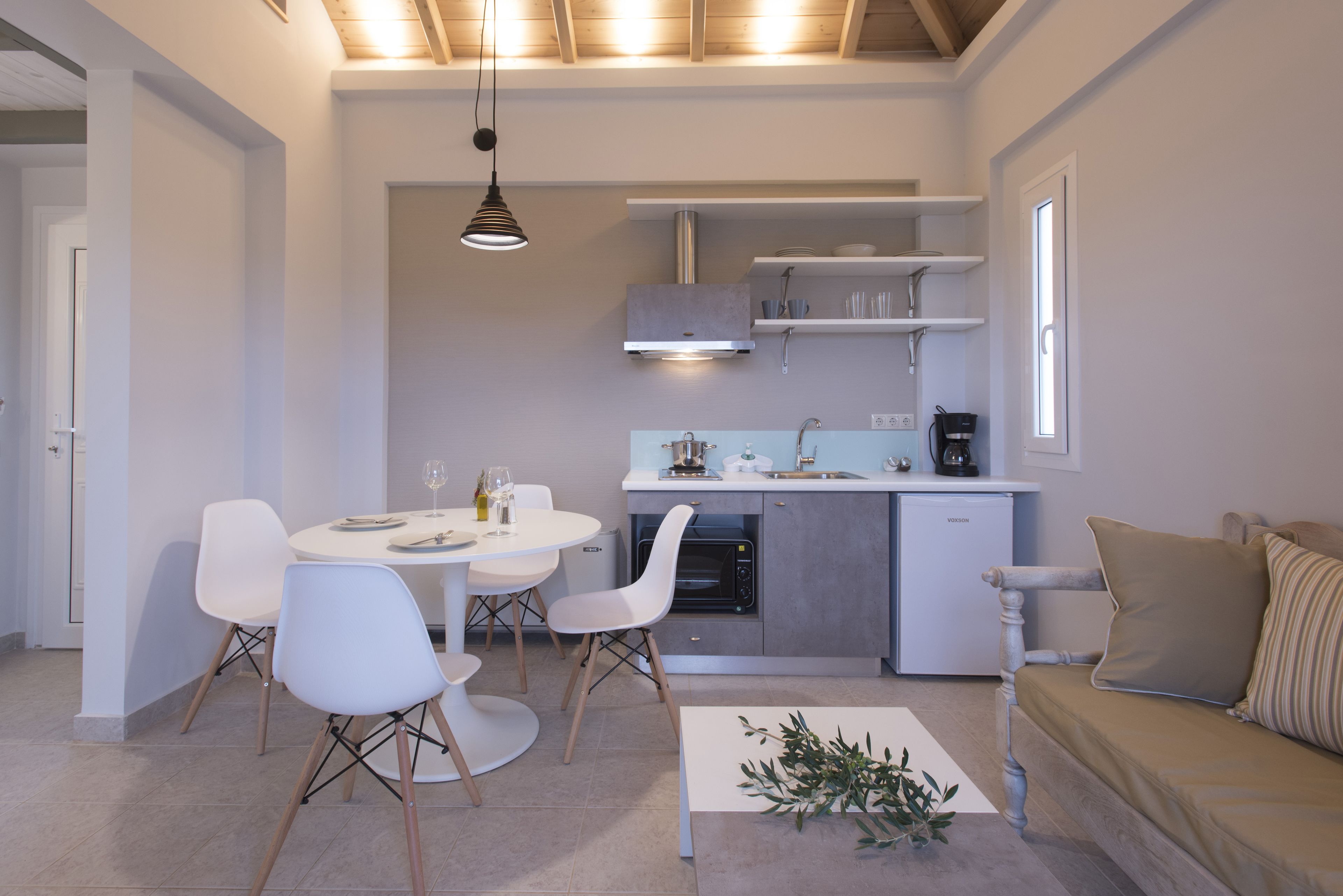 Cozinha moderna com armários brancos, forno embutido e máquina de café. Há uma sala de jantar com mesa redonda e cadeiras brancas.