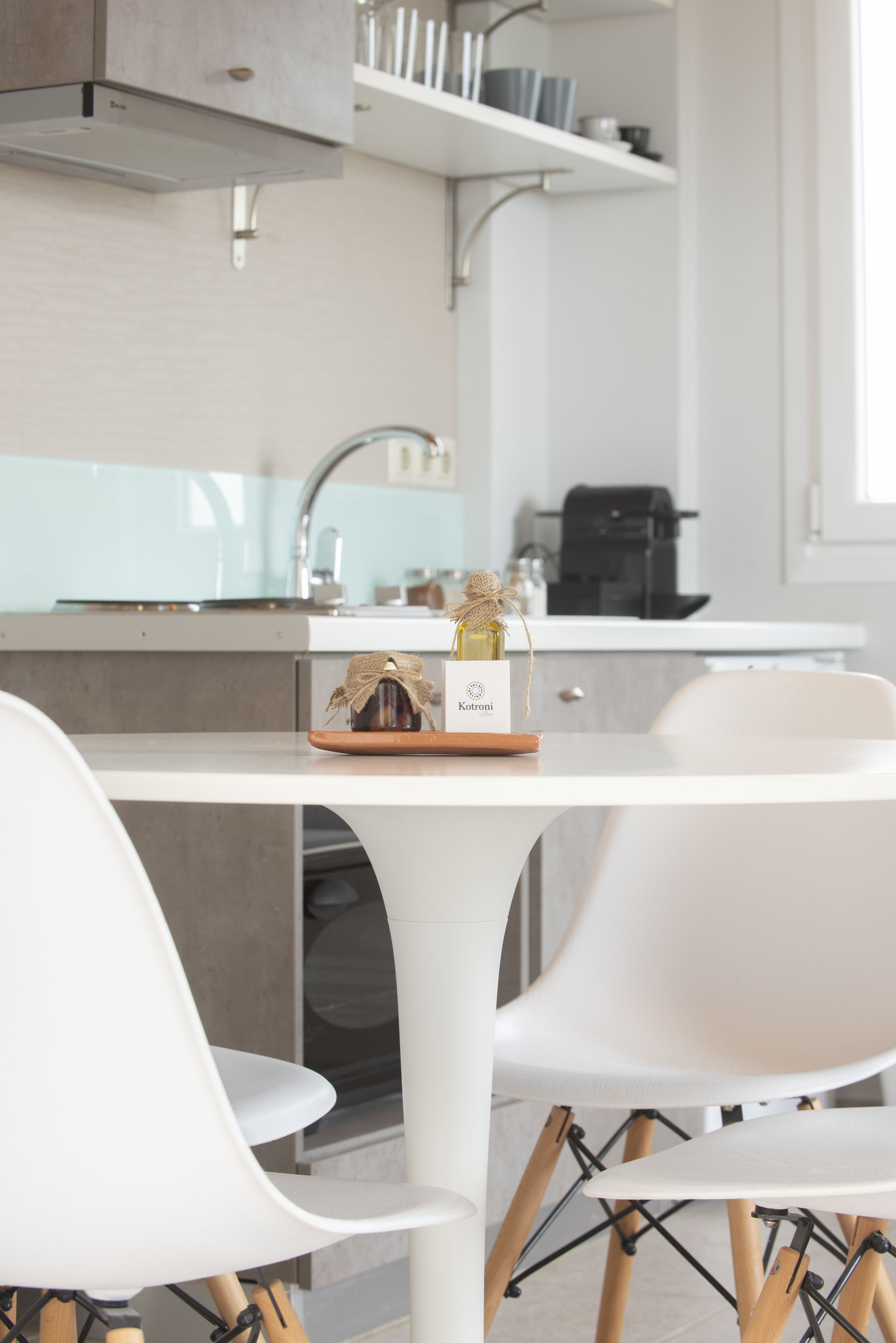 Une cuisine moderne avec des meubles blancs, un évier en acier inoxydable et une machine à café. On y trouve une petite table avec une bouteille d’huile d’olive et un pot de miel.