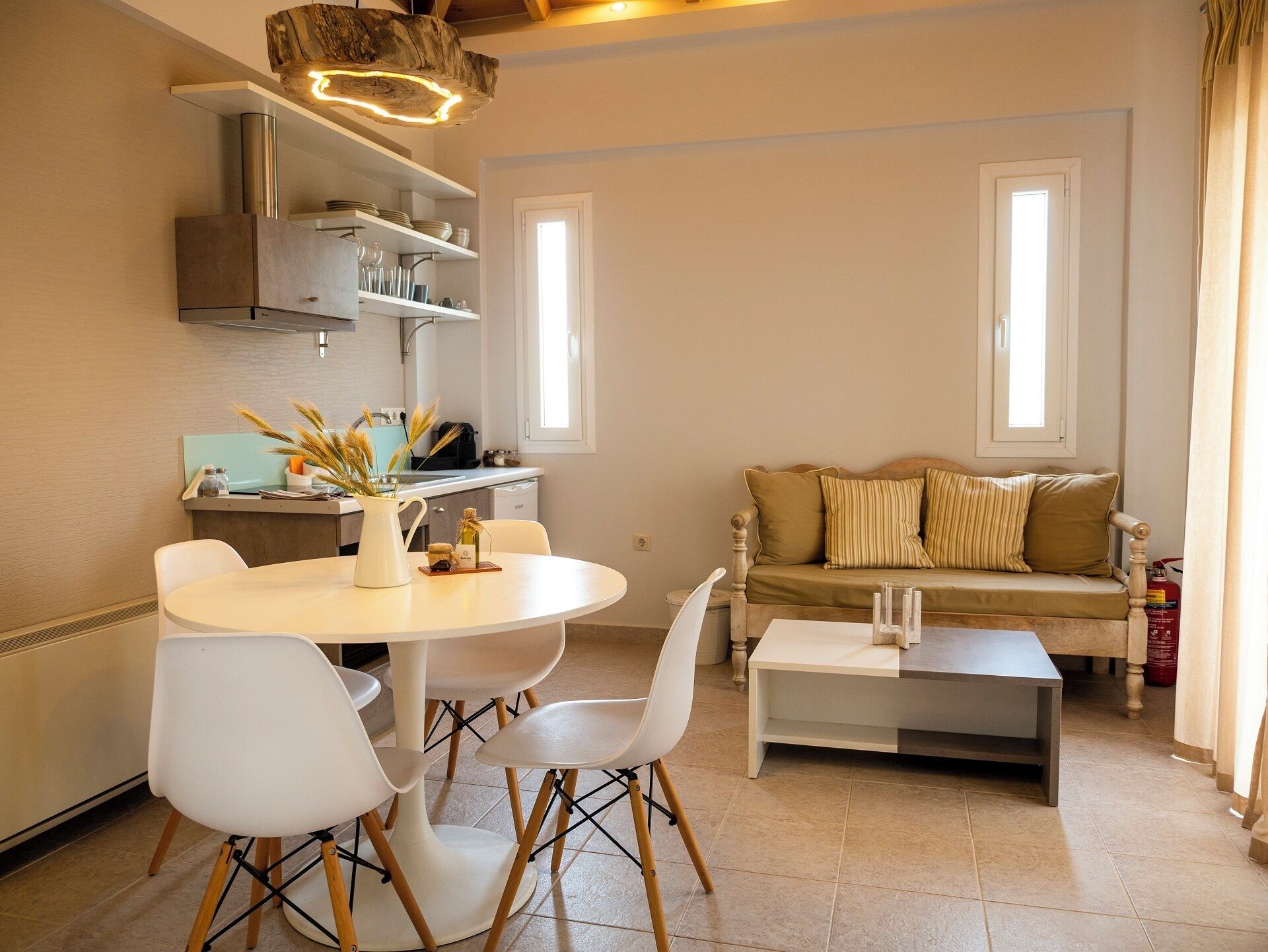 A modern living room with a round dining table, white chairs, a sofa with striped cushions, and a coffee table. There is a kitchen area with a stove, shelves, and a hanging light fixture.