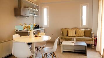 A modern living room with a round dining table, white chairs, a sofa with striped cushions, and a coffee table. There is a kitchen area with a stove, shelves, and a hanging light fixture.