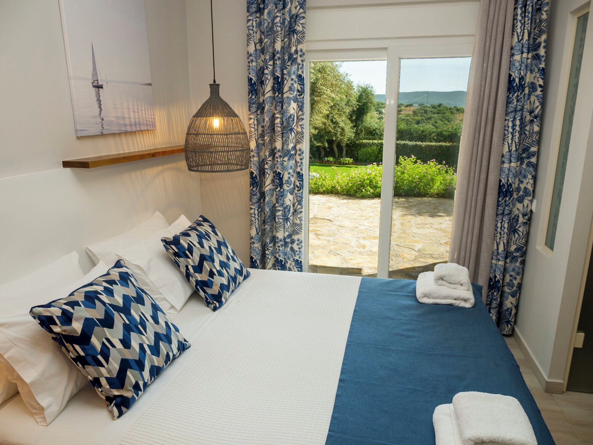 Une chambre à coucher avec un lit, des oreillers, un luminaire suspendu et une vue sur un jardin, visible à travers une porte vitrée.