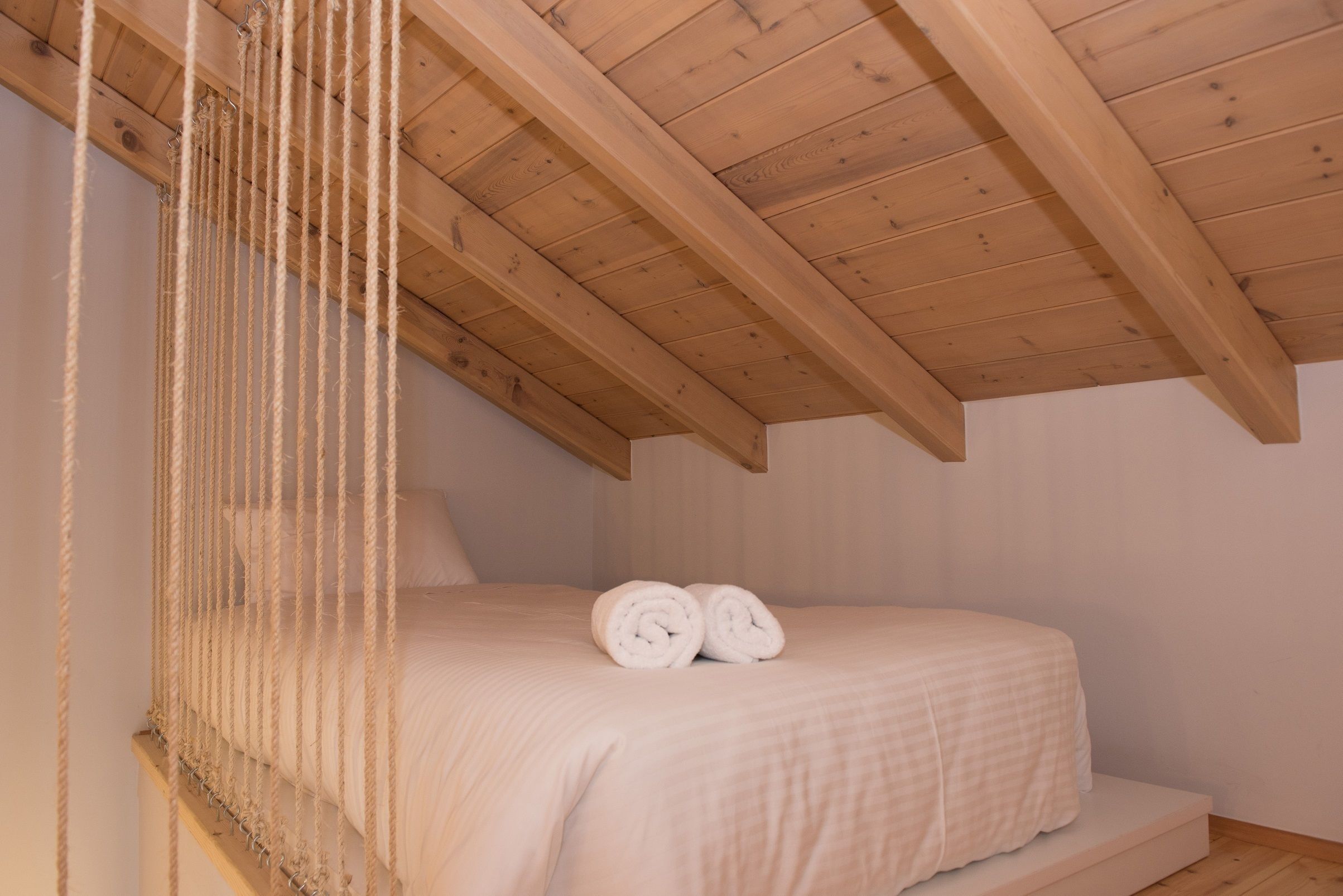 A bedroom with a wooden ceiling, a bed with white bedding, and a rope ladder.