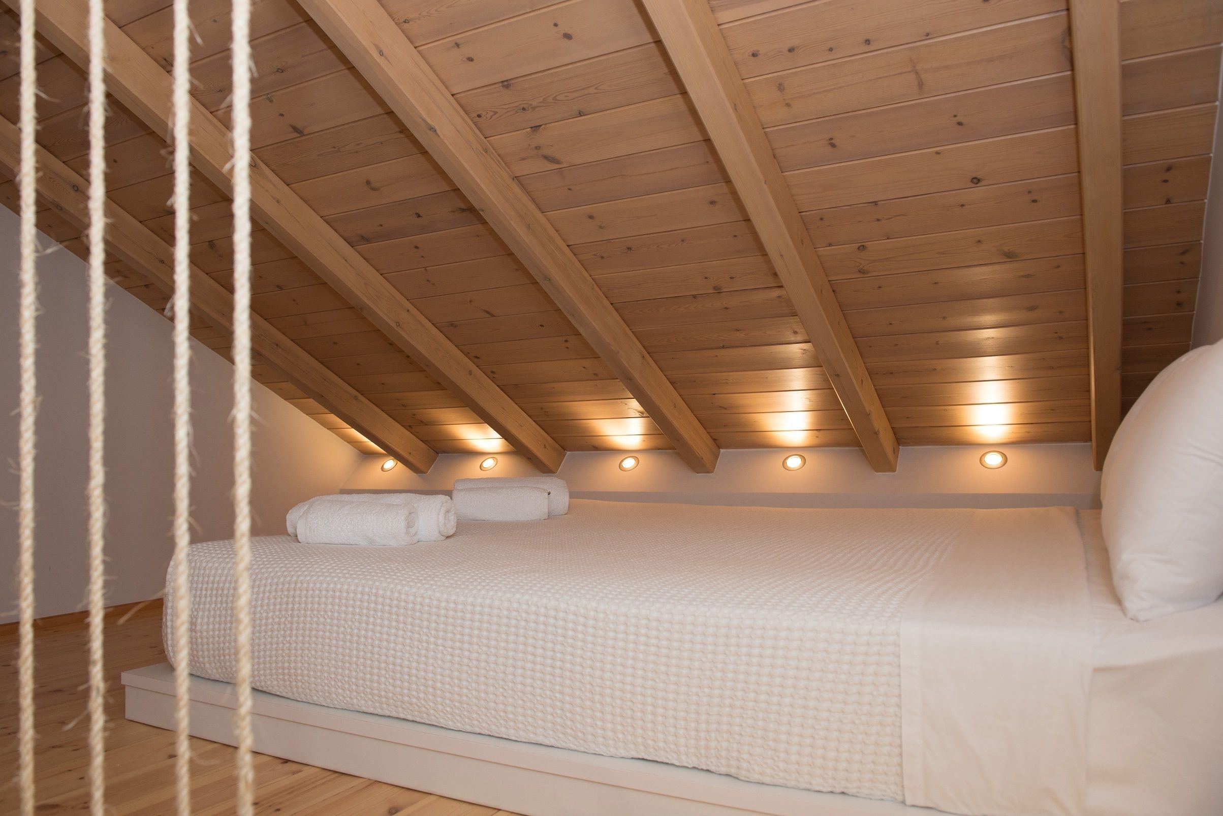 Un dormitorio con techo inclinado, vigas de madera y una cama con ropa blanca.
