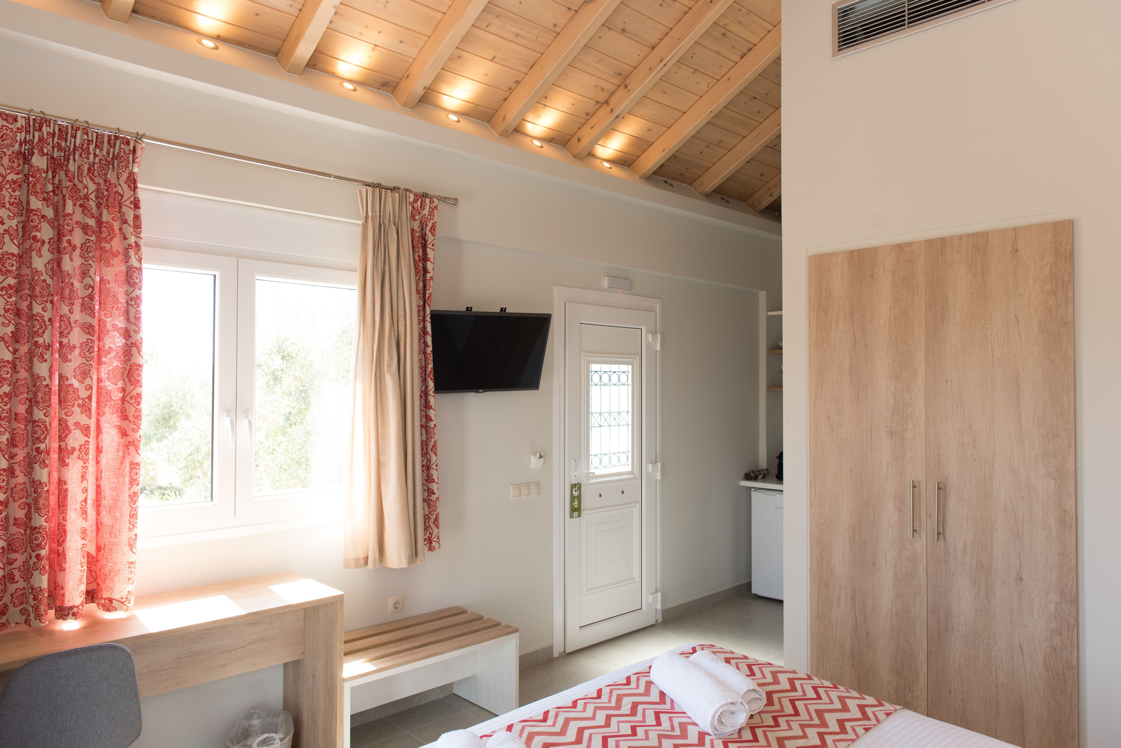 A bedroom with a wooden ceiling, a bed with a red and white patterned bedspread, a wooden bench, a desk, a TV mounted on the wall, and a wardrobe.