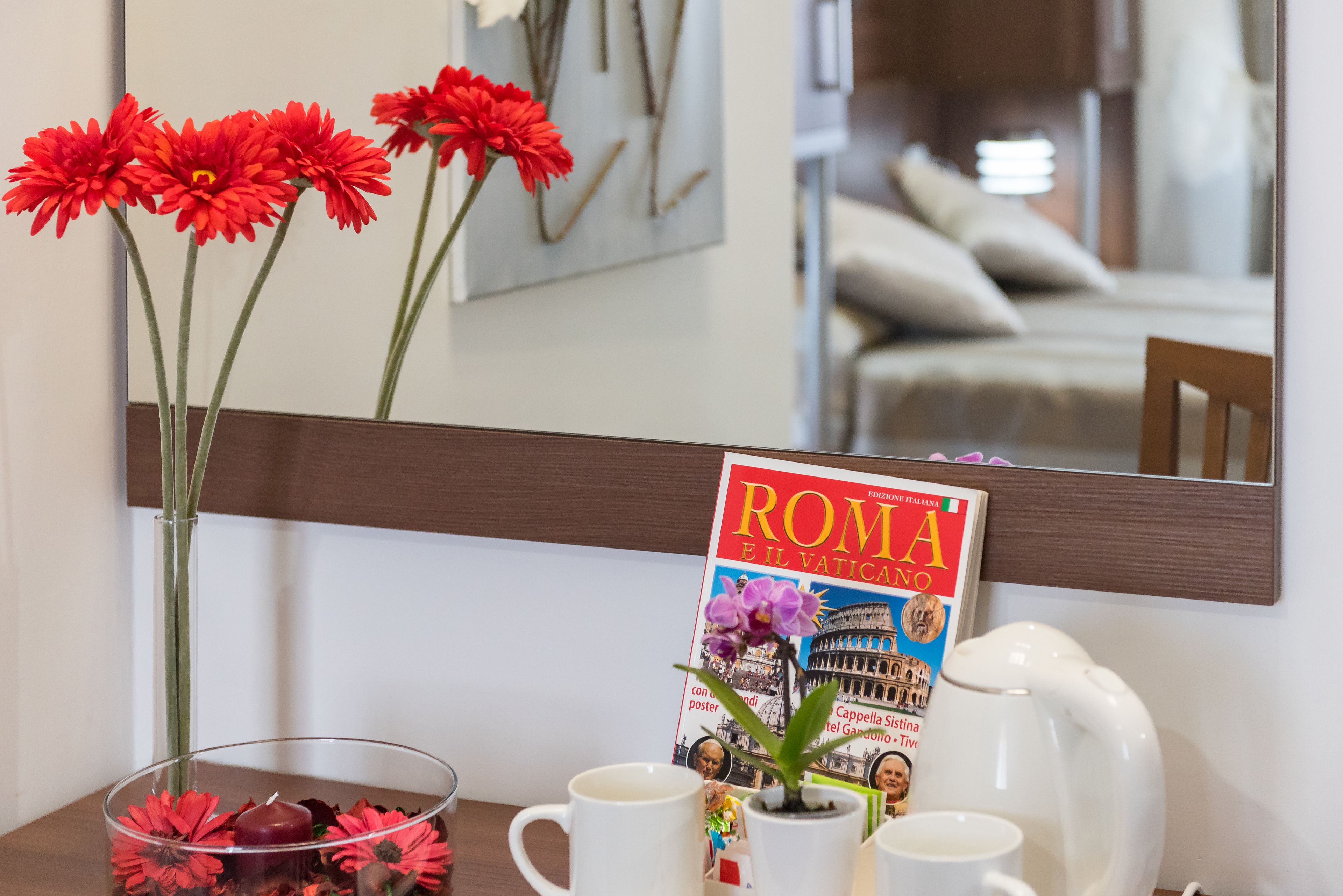 Habitación con un atril que sostiene la revista 'ROMA E IL VATICANO', un florero con flores rojas, una cafetera y un espejo que refleja la habitación.