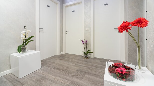 Um corredor com três portas brancas, uma mesa consola branca com um vaso de flores e um vaso de vidro com flores vermelhas sobre um pedestal branco.