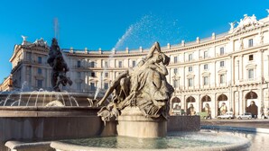 Uma fonte imponente com diversas esculturas, em frente a um amplo edifício clássico com várias arcos e colunas.