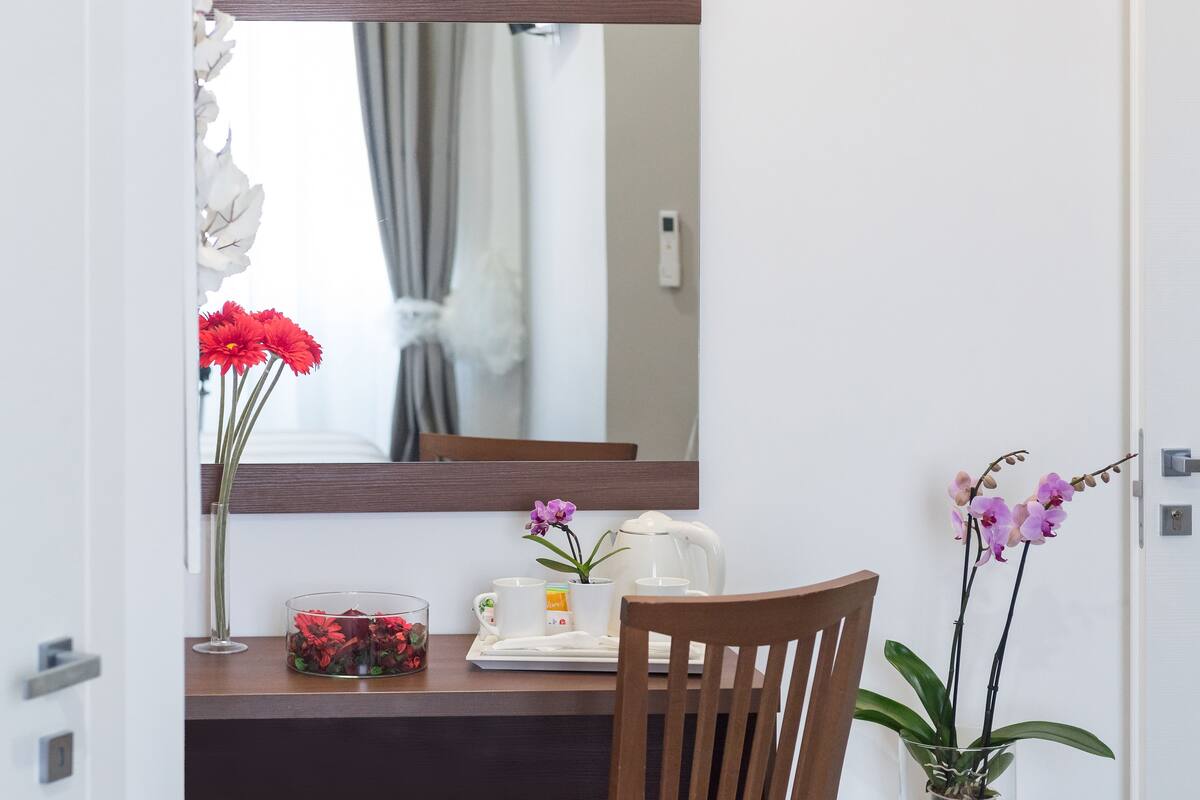 A hotel room with a wooden chair, a small table with a white kettle, a vase with red flowers, and a mirror reflecting a window with curtains.