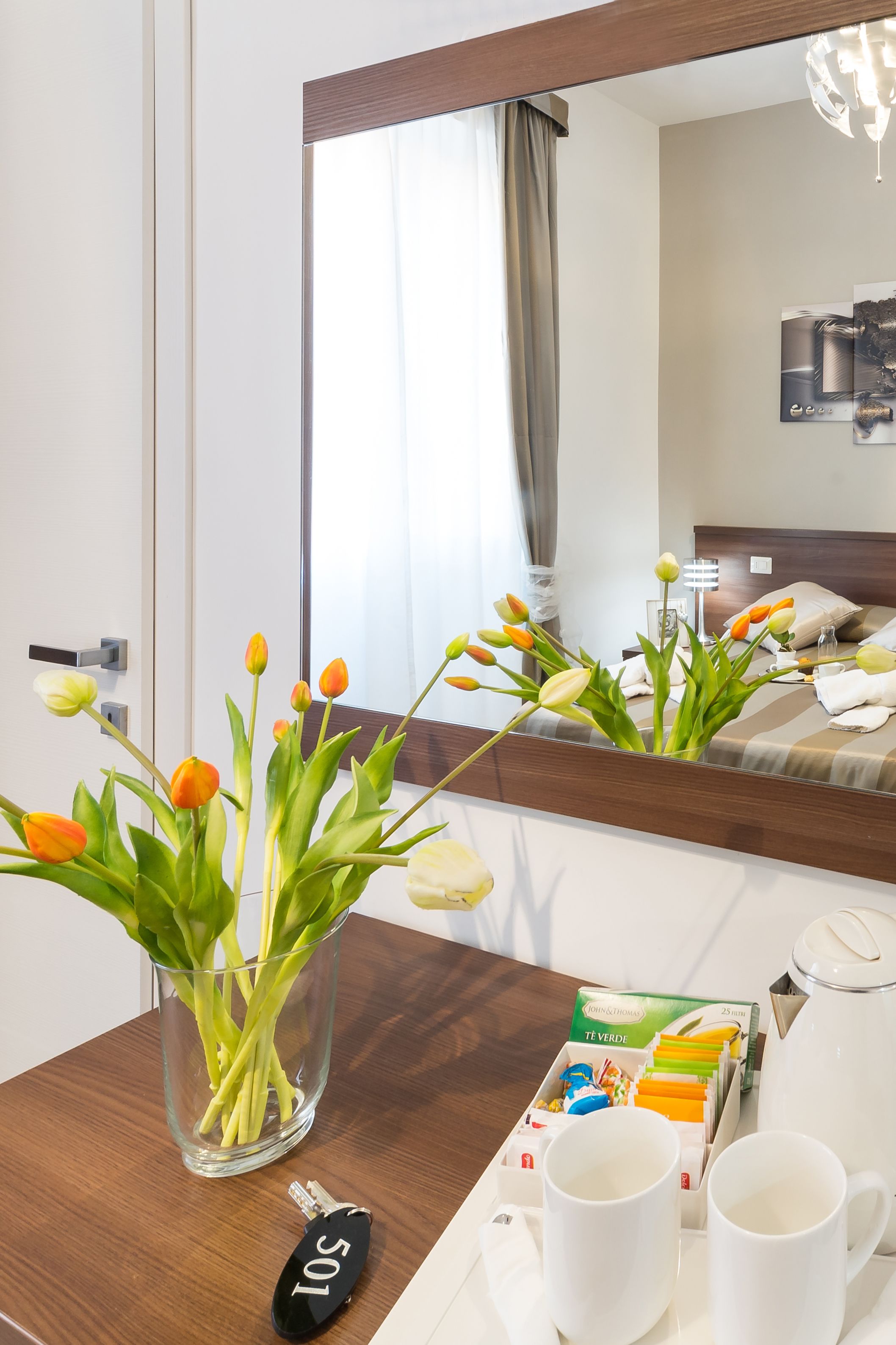 A hotel room with a wooden bedside table, a vase of tulips, a key, a tray with two white mugs, and a box of tea bags.
