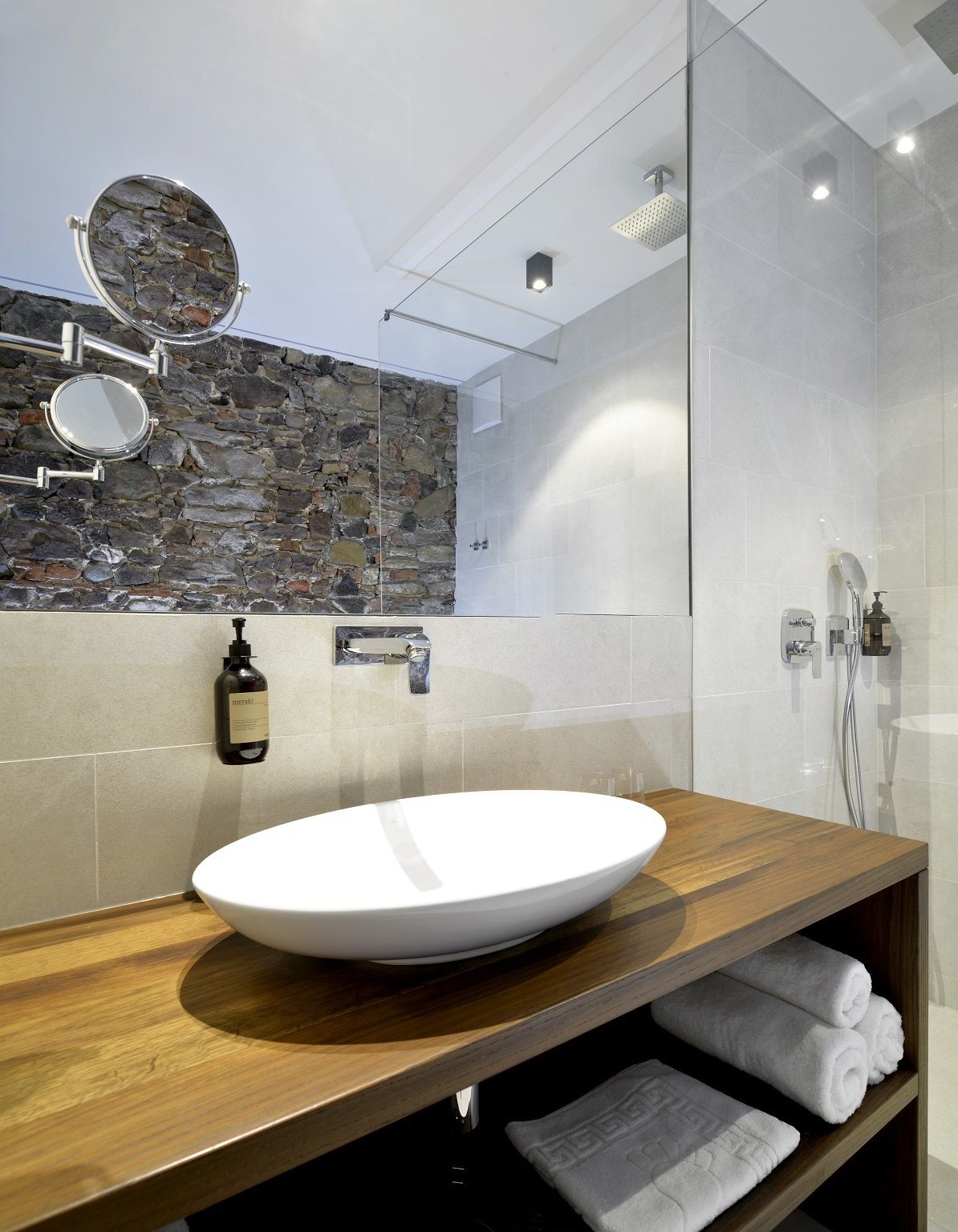 A modern bathroom with a stone wall, a large mirror, a white sink, and a shower area.