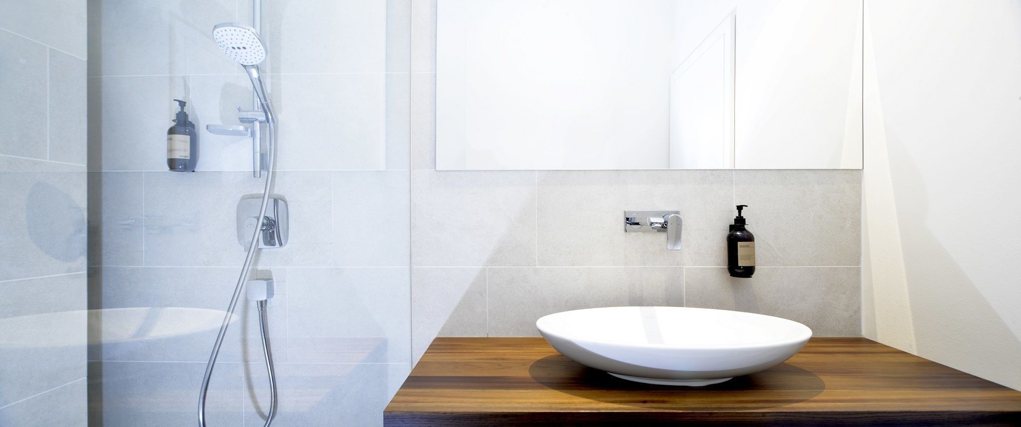 Un baño moderno con un lavamanos blanco, una encimera de madera, una ducha con cabezal de ducha manual y una botella de champú en la pared.