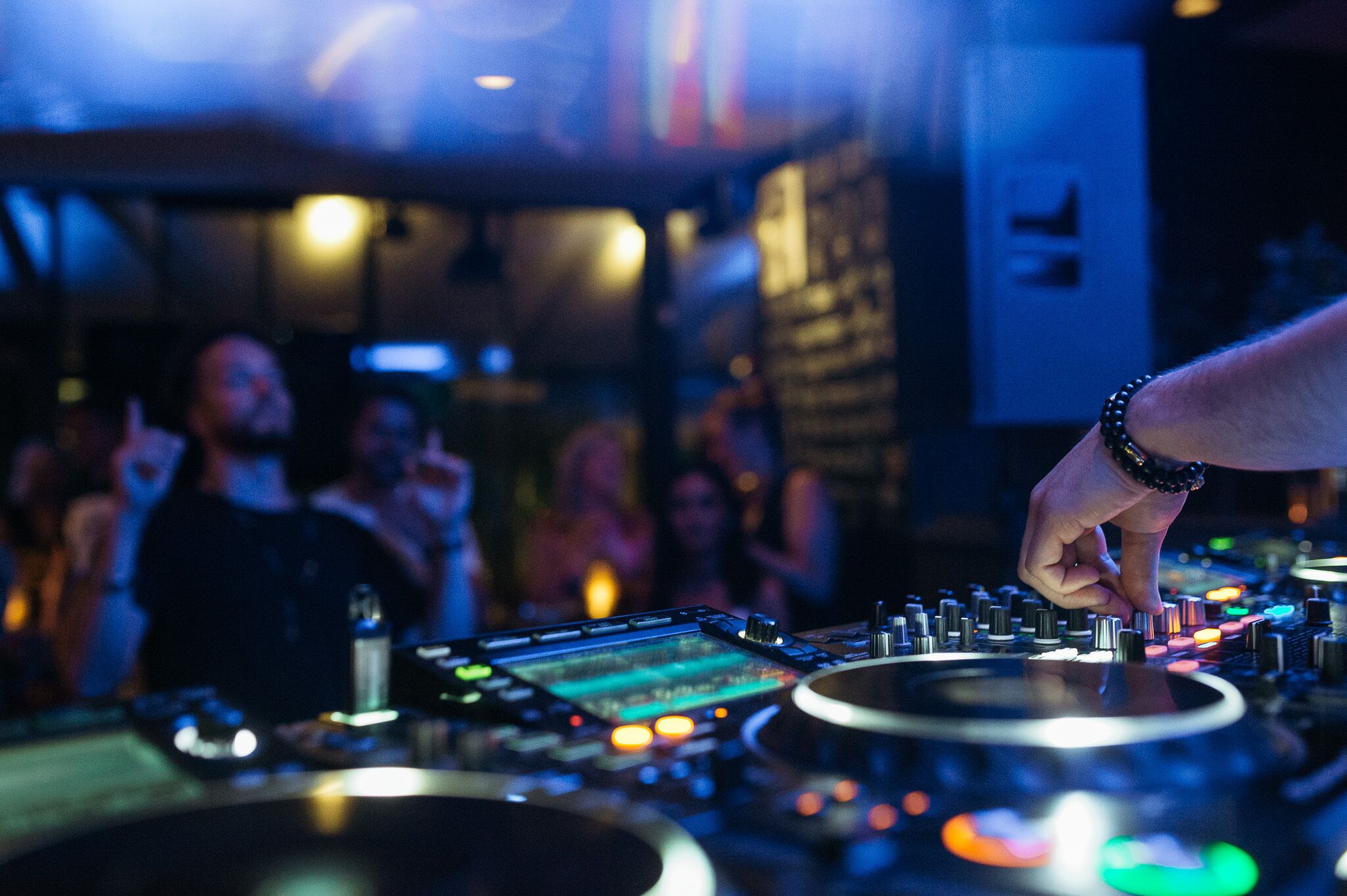 A DJ booth with a hand adjusting equipment, surrounded by a crowd in a dimly lit venue.