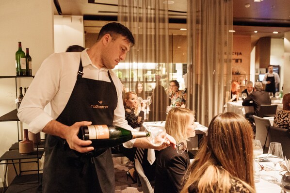 Ein Restaurantinneres mit gedeckten Tischen, einem Kellner, der eine Champagnerflasche serviert, und einer Bar mit Flaschen.