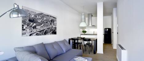 A modern living room with a grey sofa, a dining table with chairs, a black refrigerator, and a kitchen area with a hanging light fixture.