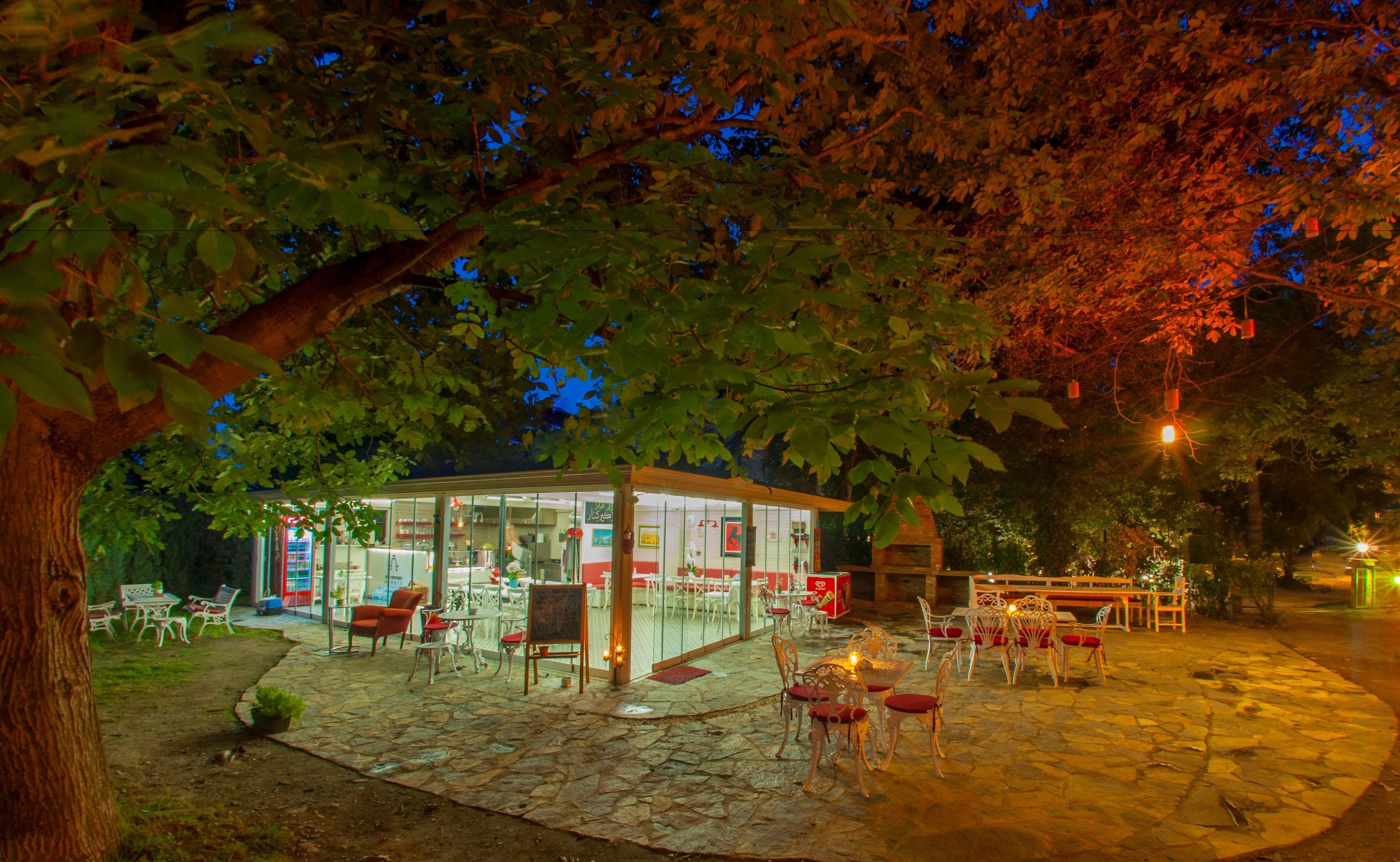Un café en plein air doté d’un sol en pierre, de tables et de chaises, entouré d’arbres et éclairé par un éclairage d’ambiance.