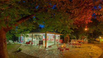 Un café en plein air doté d’un sol en pierre, de tables et de chaises, entouré d’arbres et éclairé par un éclairage d’ambiance.