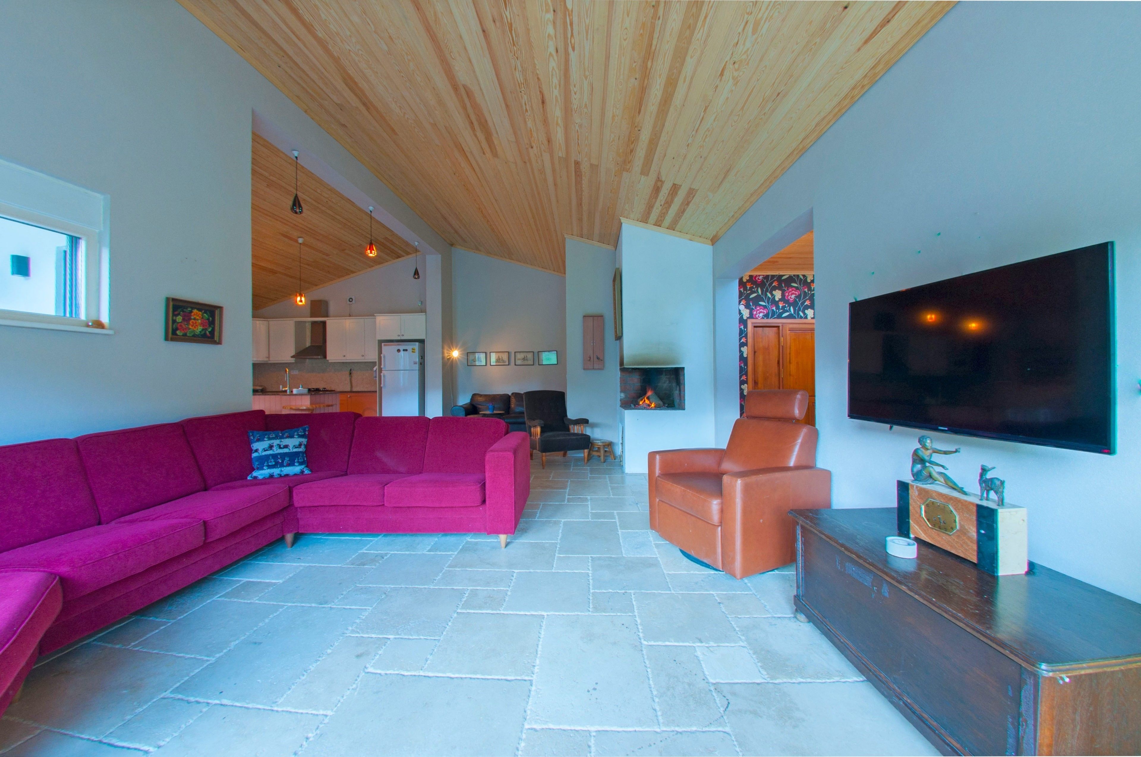 A spacious living area with a wooden ceiling, a large flat-screen TV, a pink sectional sofa, a brown leather armchair, and a stone-tiled floor.