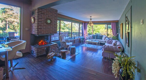A cozy living room with a brick fireplace, comfortable seating, and large windows offering a view of the outdoors. - Villa Kirkpinar Sapanca (Sapanca)