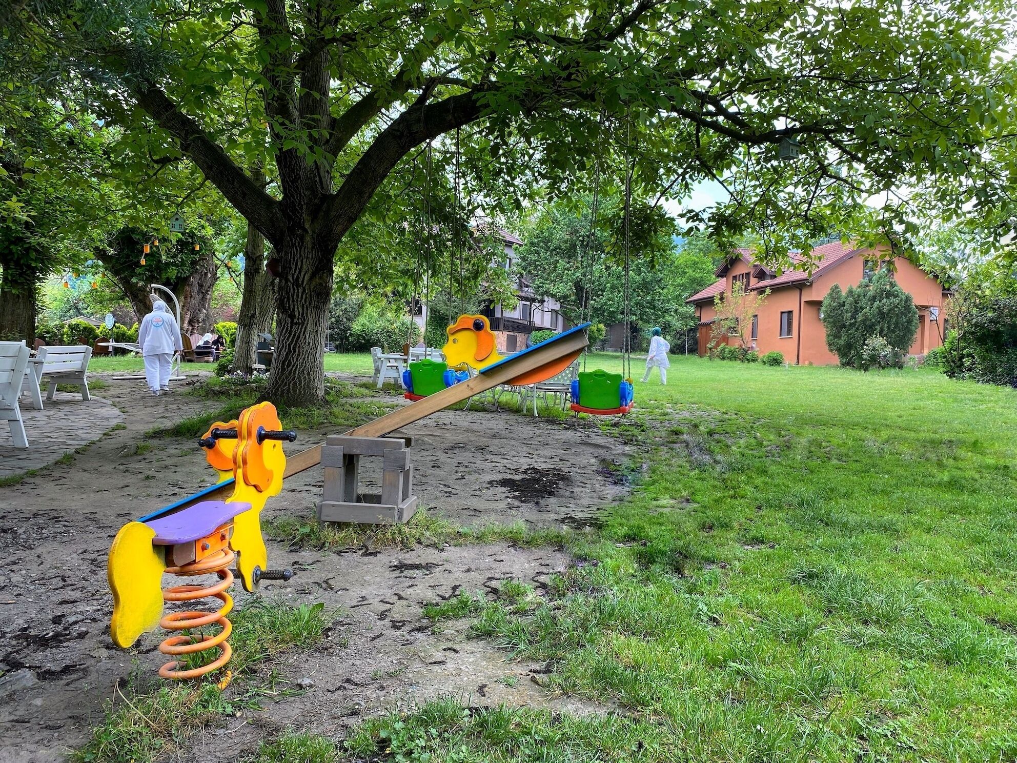 A playground with seesaws and swings, surrounded by trees and a grassy area.