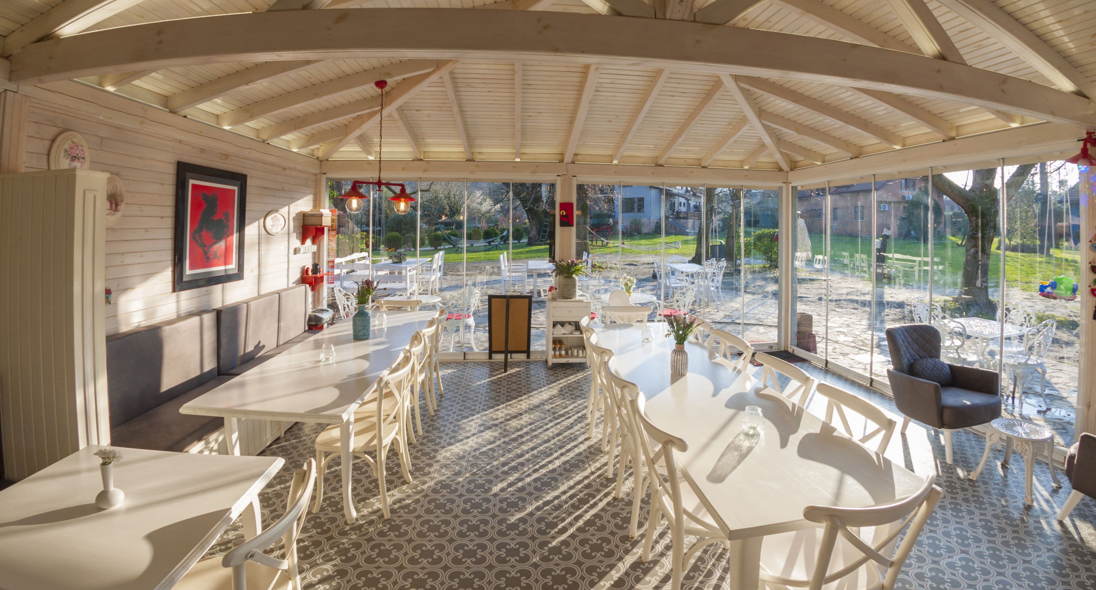 A covered outdoor dining area with white furniture, a patterned floor, and a view of a garden and houses in the background.