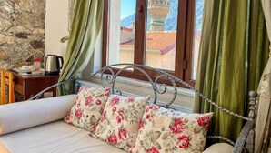 A room with a stone wall, a bed with floral pillows, a view of a minaret, and a window with green curtains. - Fulya Pension (Egirdir)