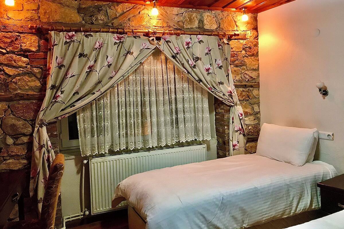 Un dormitorio con pared de piedra, cama con ropa blanca, radiador y cortinas con estampado floral.