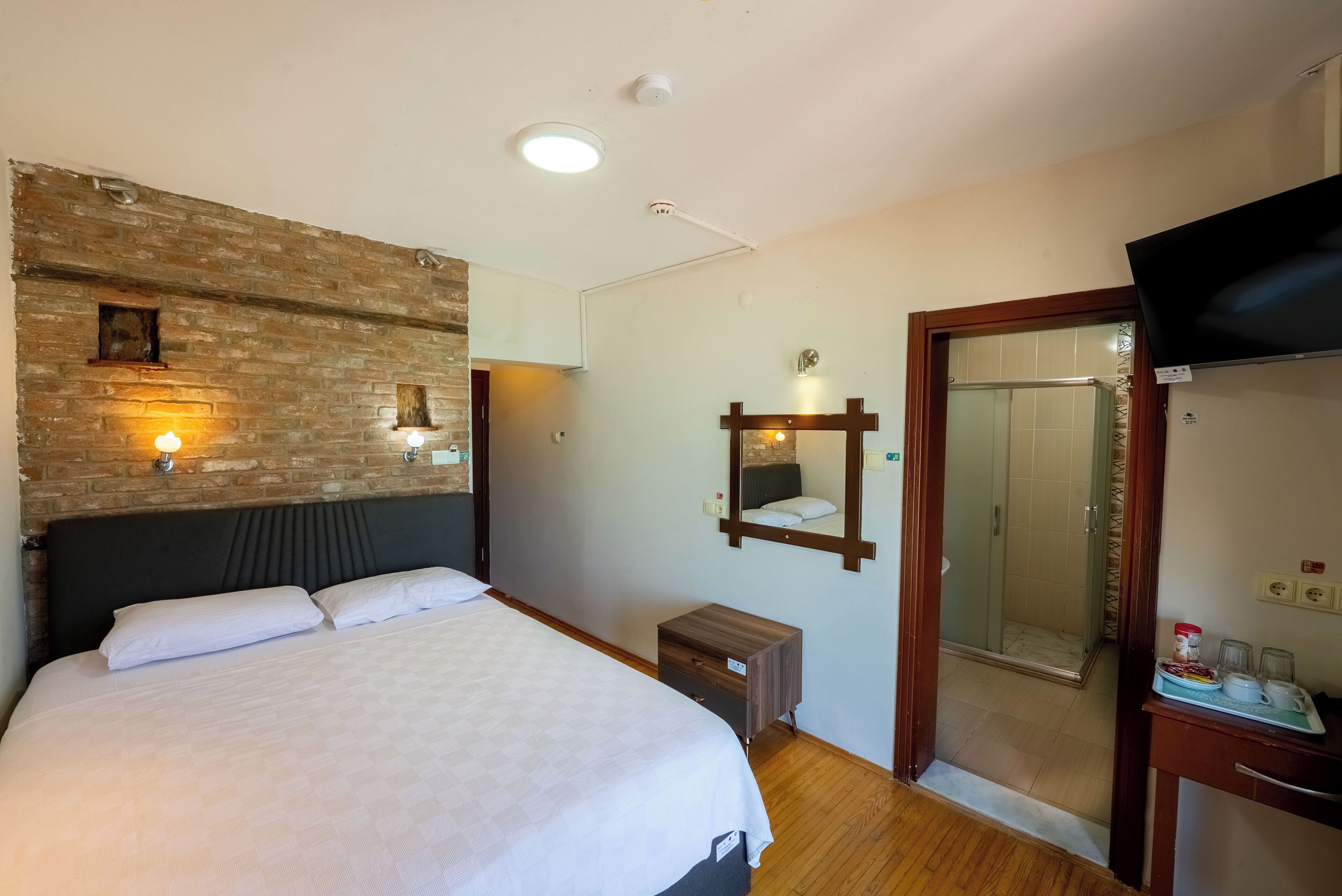 Une chambre d’hôtel avec un grand lit, une tête de lit en bois, un mur décoratif en briques, un miroir, une petite table de chevet, une télévision et un espace douche.