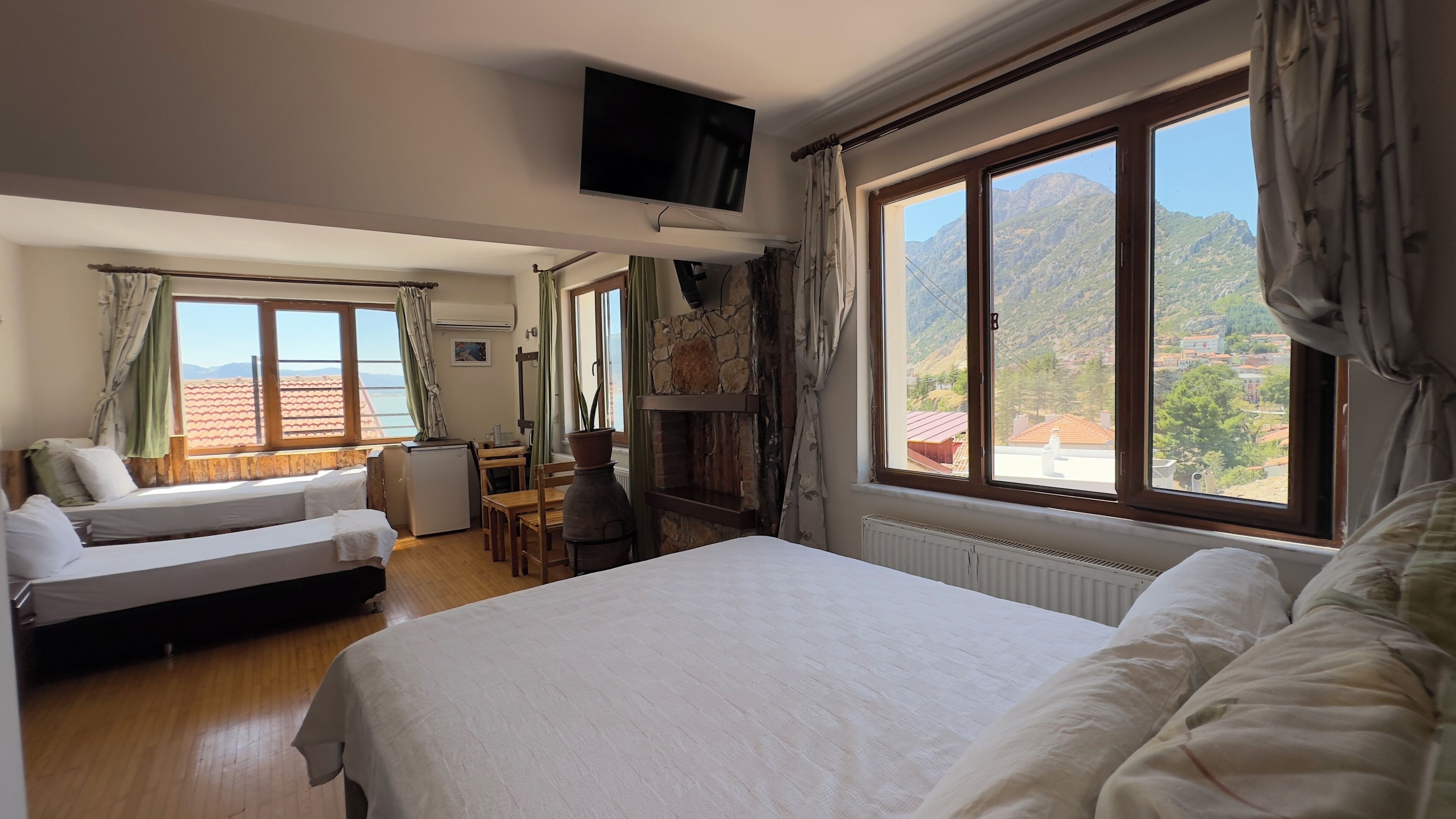 Habitación de hotel con cama, escritorio, silla, televisor y ventanas amplias con vistas a la montaña.