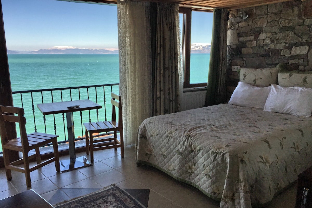 Un dormitorio con una cama grande, una mesa de madera y una silla. Se ve el mar a través de una ventana.