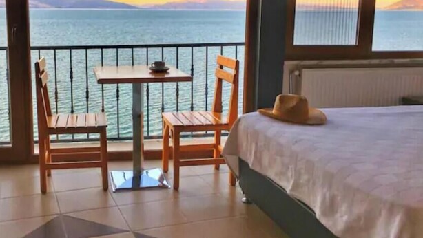 A room with a bed, a table, and two chairs facing a balcony with a view of the sea and mountains. - Fulya Pension (Egirdir)