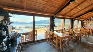 A wooden interior with tables and chairs, a kitchen area with cooking equipment, and a view of a lake and mountains through large windows. - Fulya Pension (Egirdir)