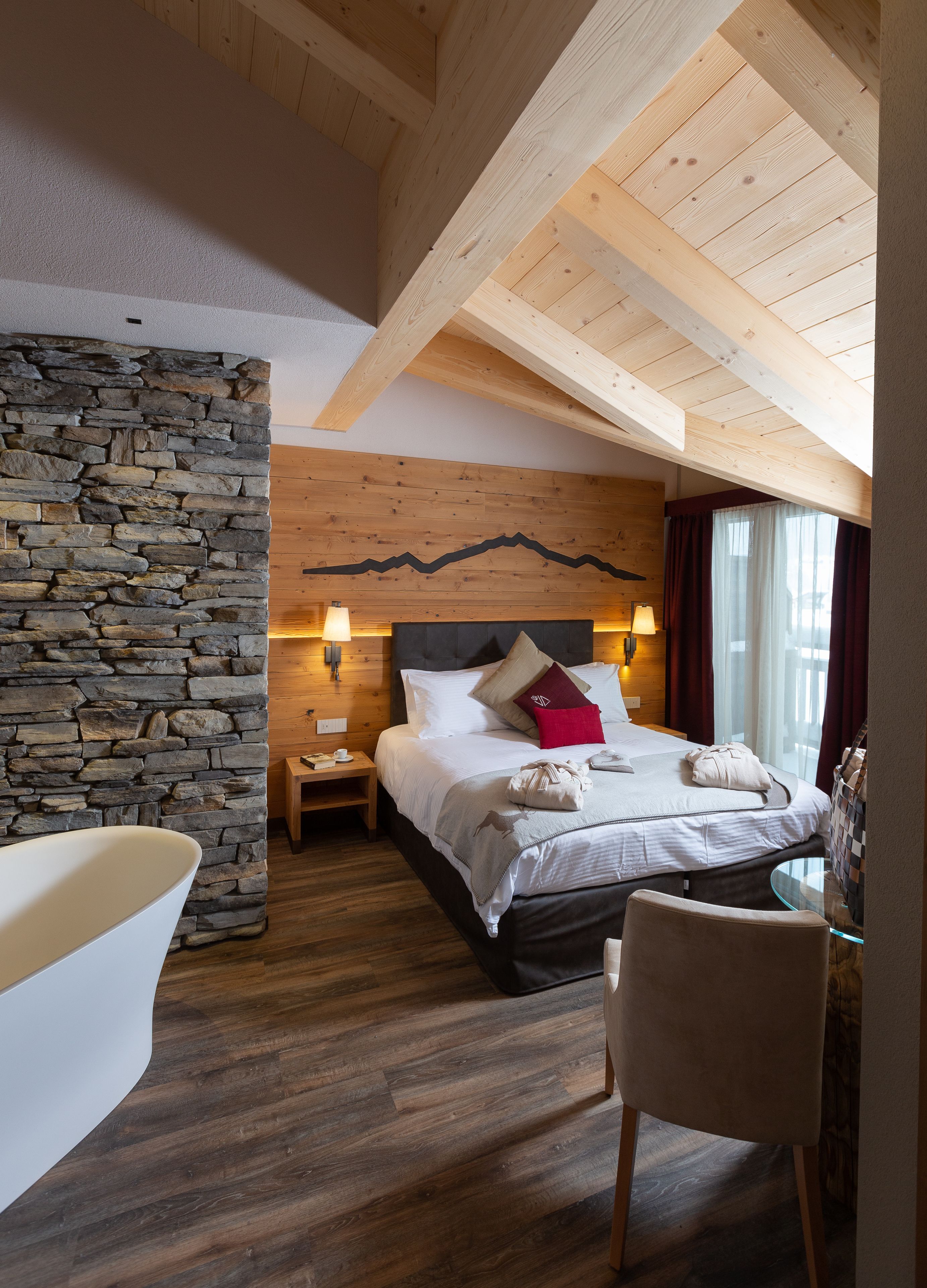 A bedroom with a stone wall, wooden ceiling beams, a bed with pillows, a nightstand, a glass table, a chair, and a bathtub.