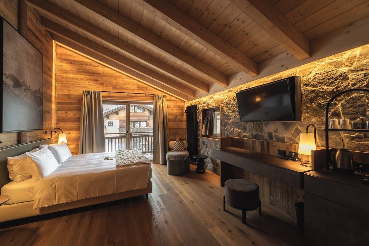 Una habitación acogedora de madera con una cama, una chimenea de piedra, un televisor montado en la pared y un balcón visible a través de un ventanal grande.