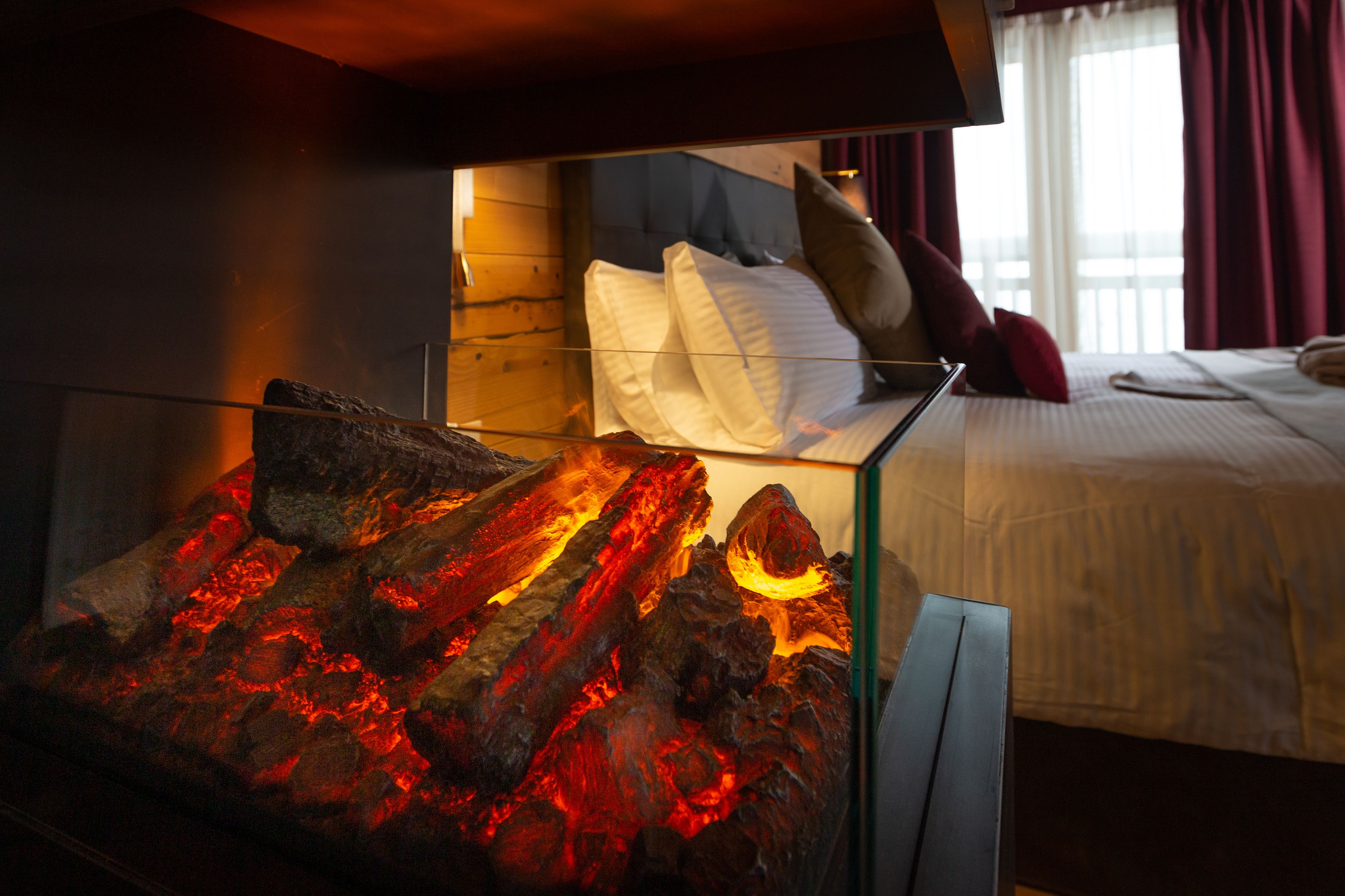 A room with a glass table displaying a lava-like display, a bed with white bedding and red pillows, and a window with curtains.